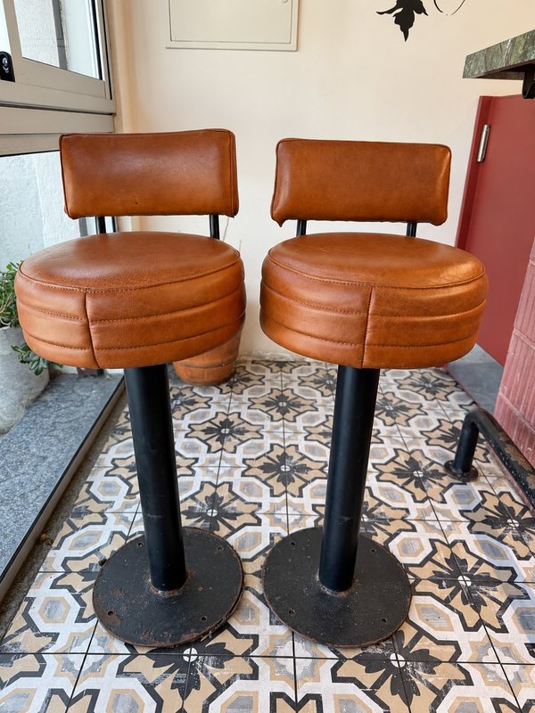 Lote #012B – Dos butacas para barra en caño grueso de amurar al piso, tapizadas en cuero color whisky – 37cm diámetro asiento, 74cm de altura al asiento y 92cm total