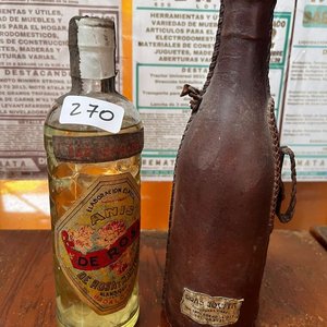 Lote siguiente: Botella de Anís Añejo De Rosas con Forro de Cuero y Tapa Tipo Porrón